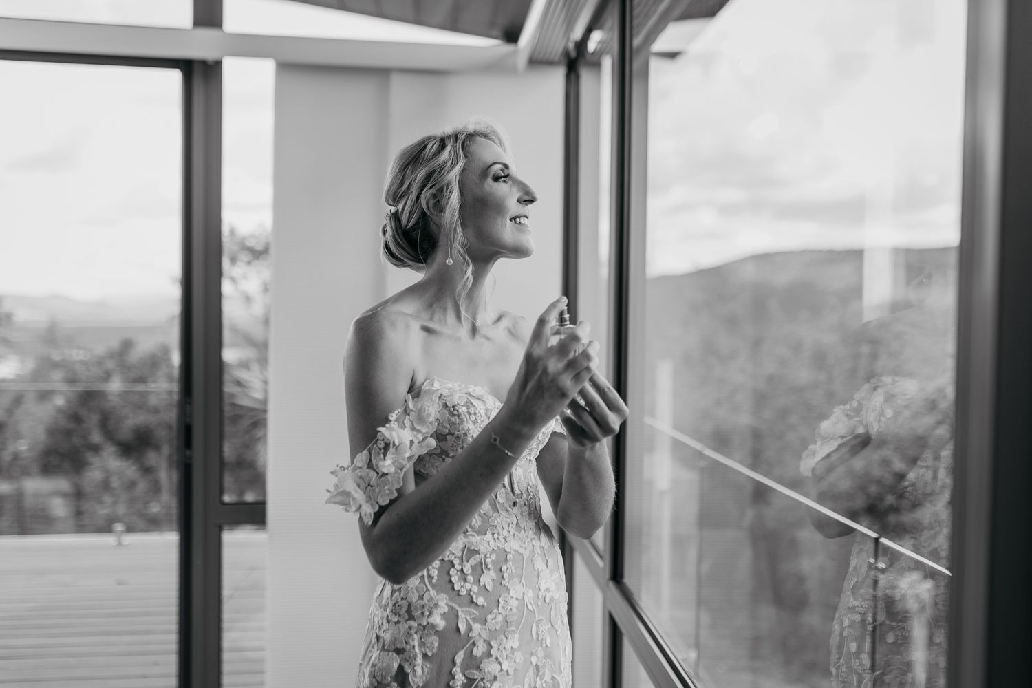 Black and white portrait of bride in lace dress near window putting on jewelry.
