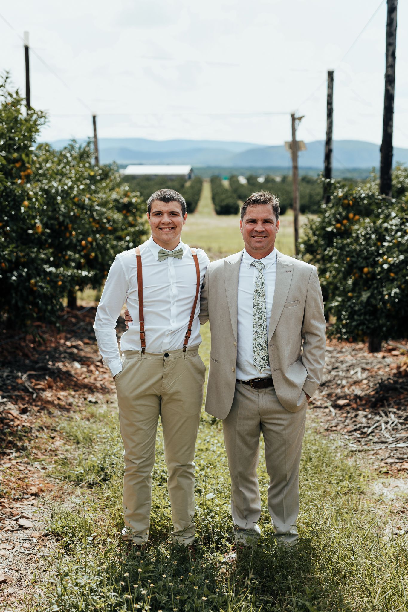 Portrait of two people in formal wear standing together in orchard with mountains in background.