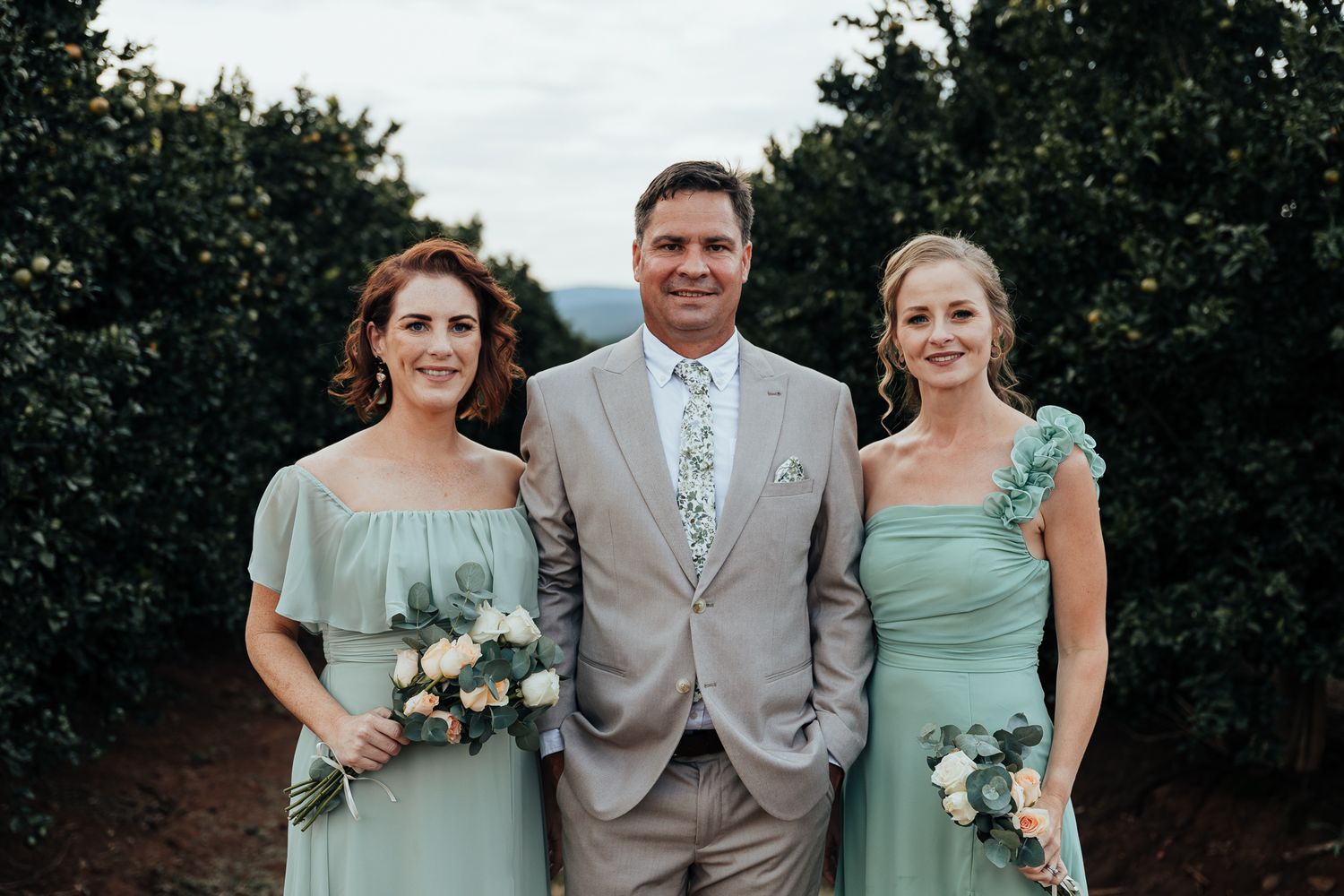 Wedding party members in mint green dresses and beige suit pose together in a garden setting.