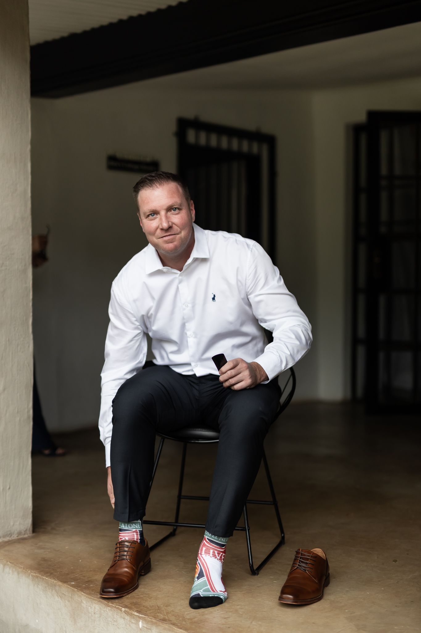 Person in white dress shirt and dark pants sitting on stool in minimalist setting.
