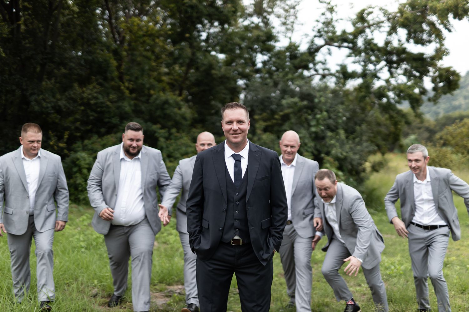 Groomsmen pose playfully behind the groom in a grassy field with trees.