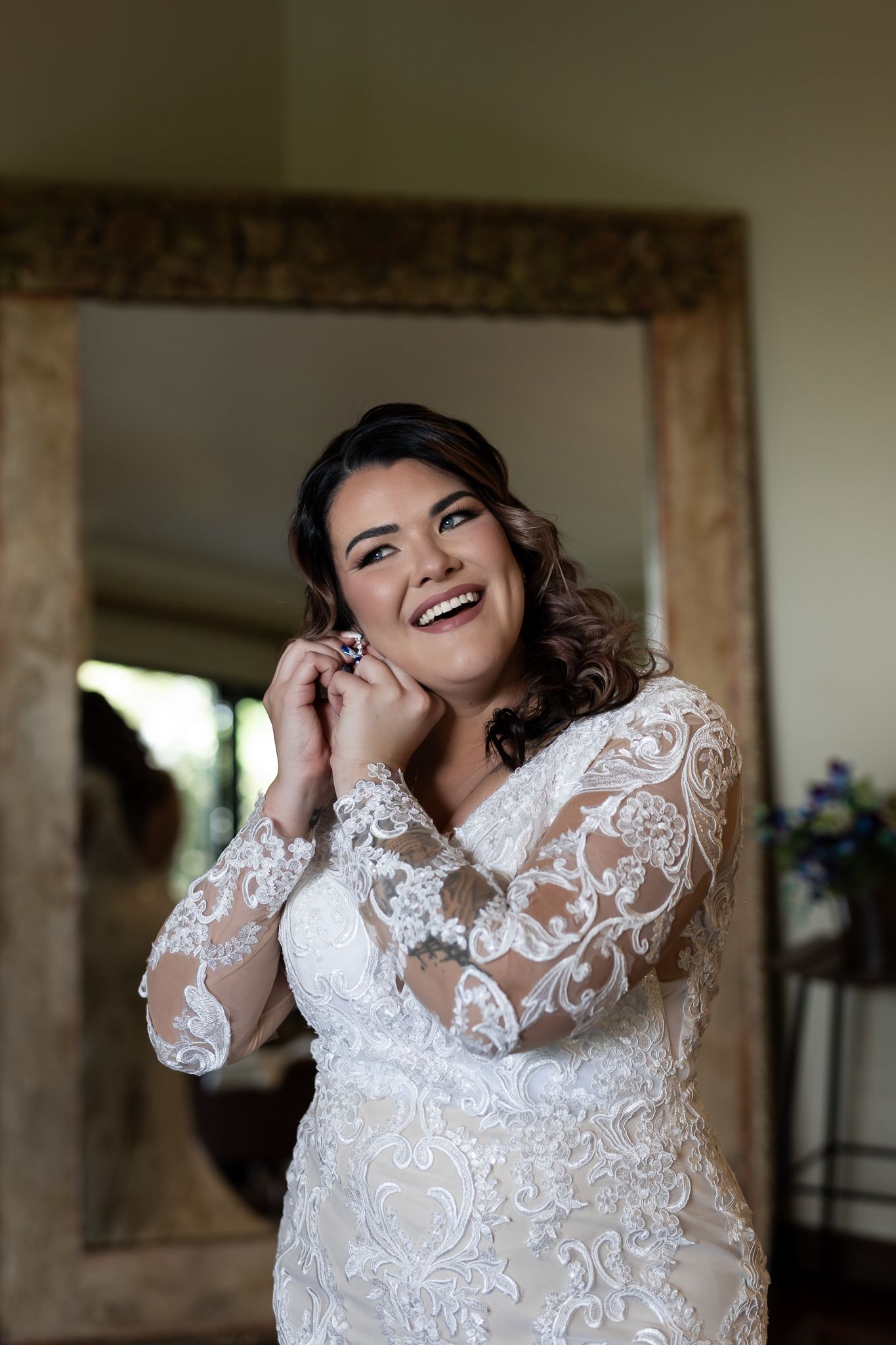 Bride wearing a lace wedding dress smiles while putting on earrings in front of rustic mirror.