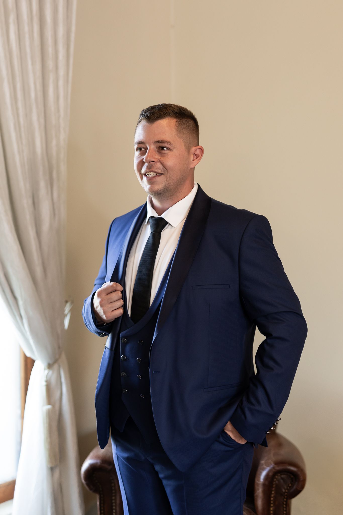 Portrait of person in navy suit smiling near window and curtains.