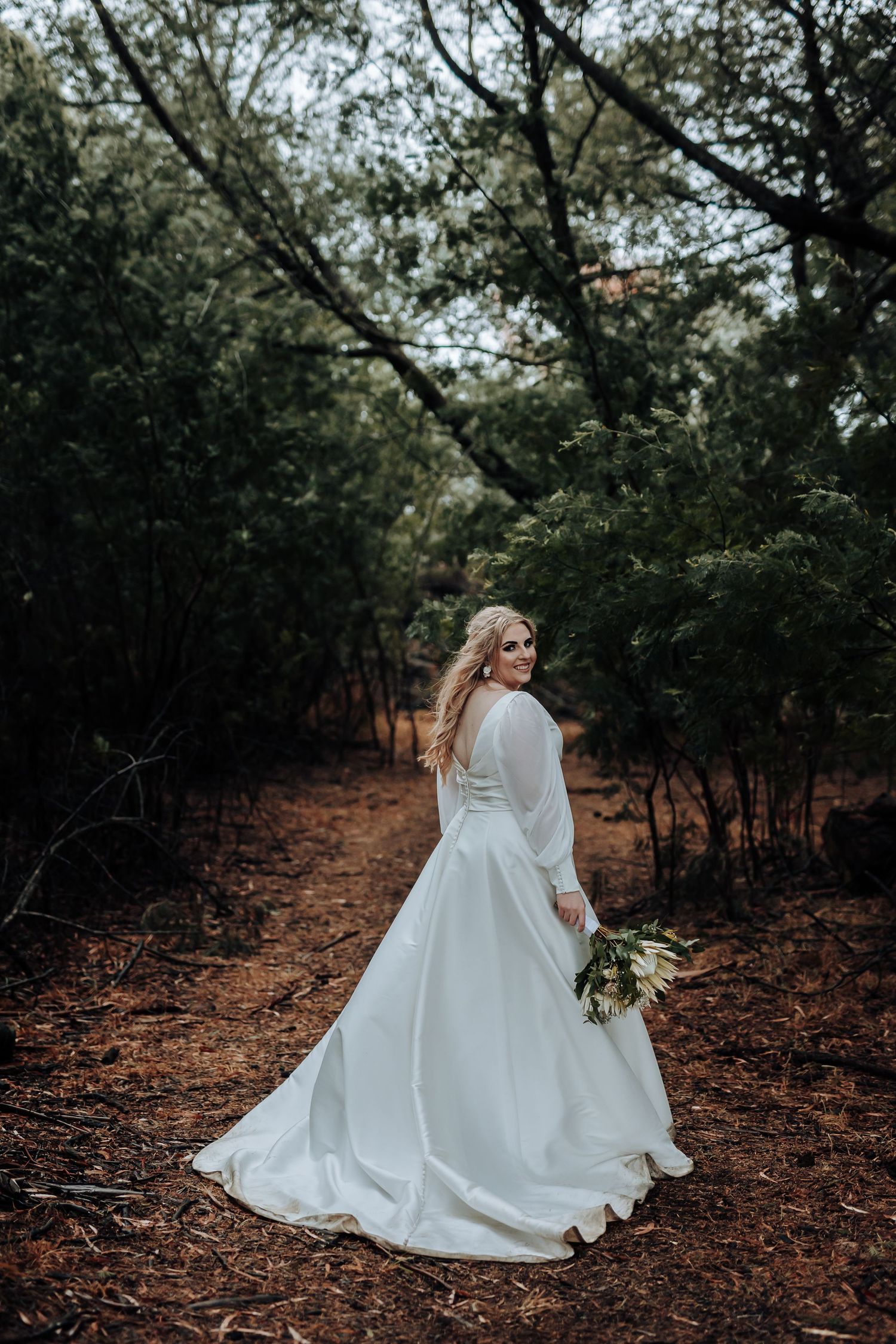 Bride in flowing white dress twirls on rustic forest path holding natural bouquet.