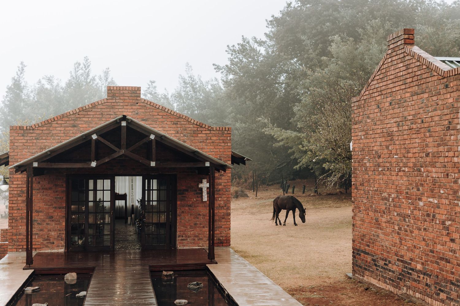 Red brick building with covered porch and horse grazing in foggy background.