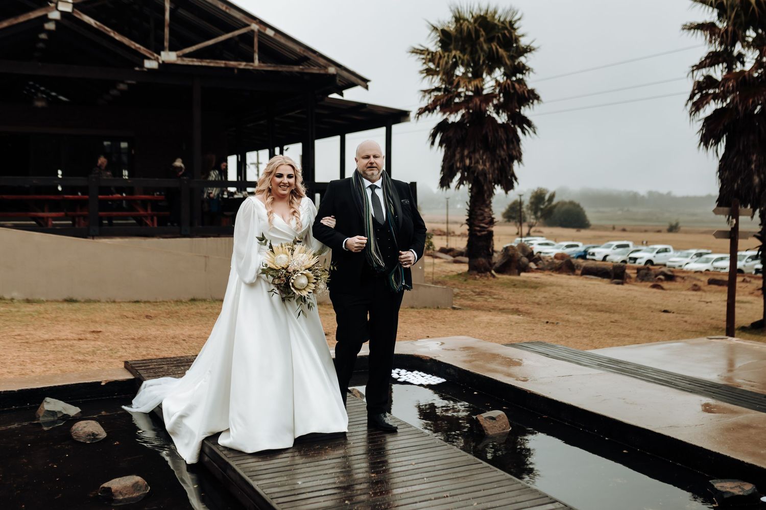 A couple walks down a wet boardwalk during their rainy outdoor wedding ceremony with palm trees in background.