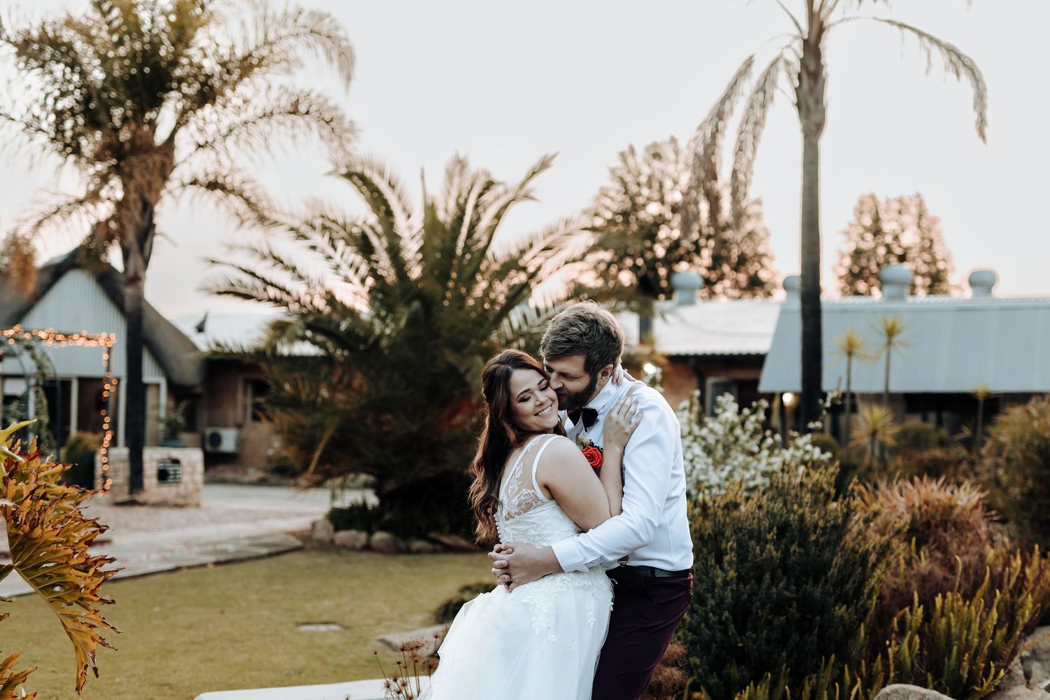 Wedding couple poses in tropical garden setting with palm trees and blue building in background.