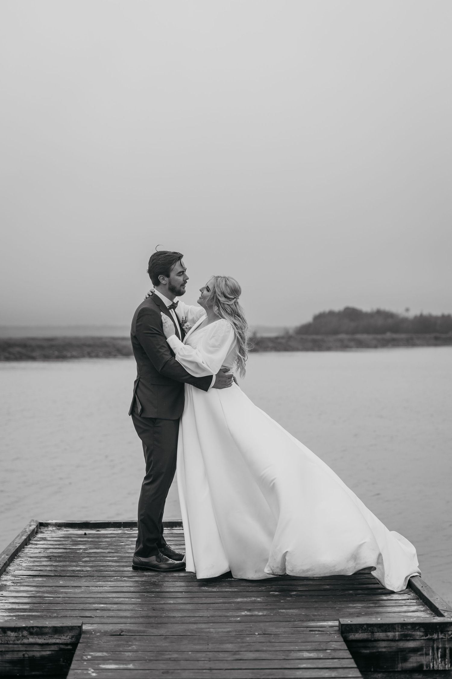Black and white portrait of couple in wedding attire embracing on waterfront dock.