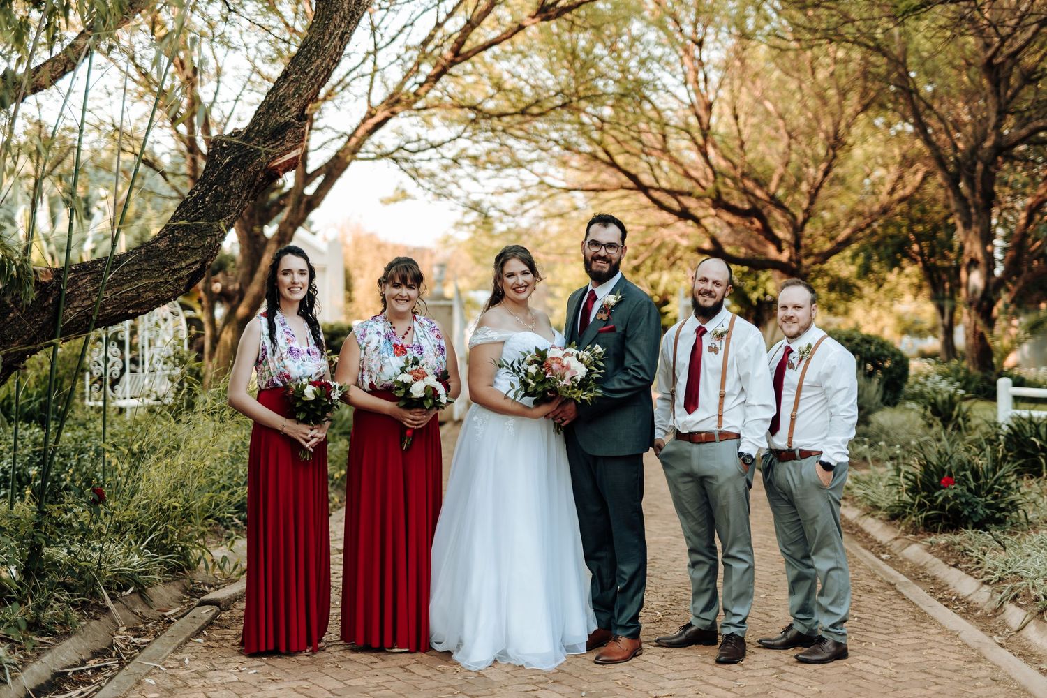 A wedding party in burgundy and grey attire poses on a tree-lined path.