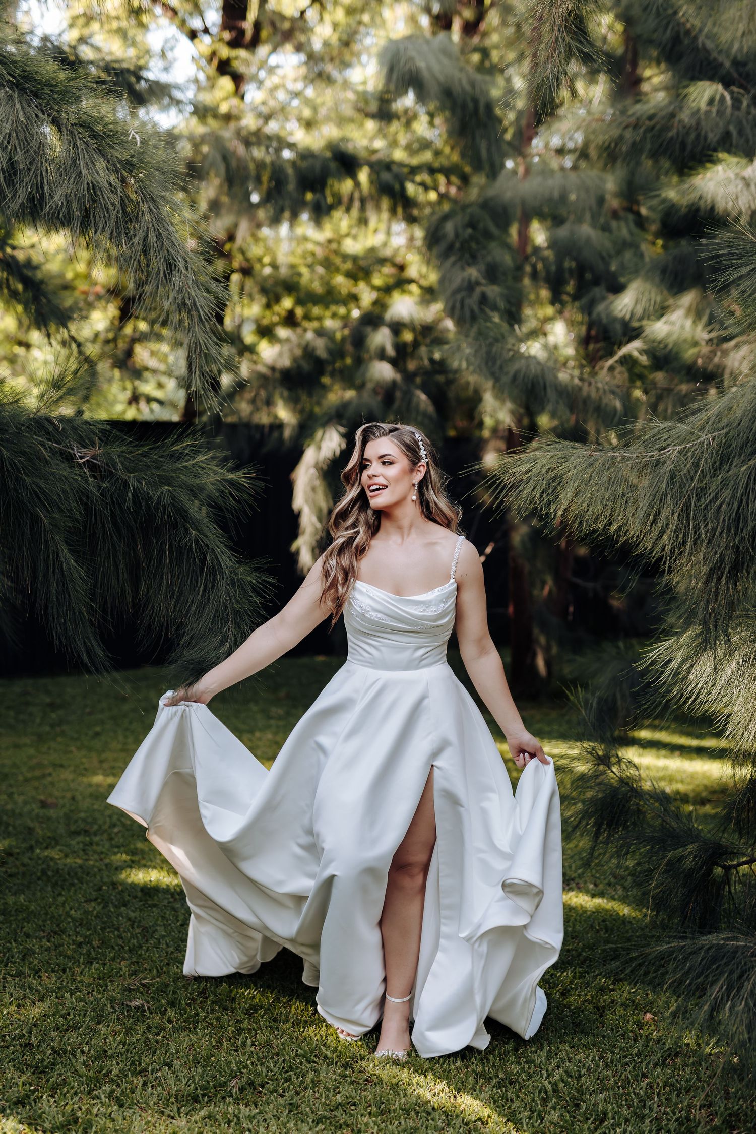 A flowing white wedding dress twirls gracefully in a sunlit garden setting with pine trees in the background.