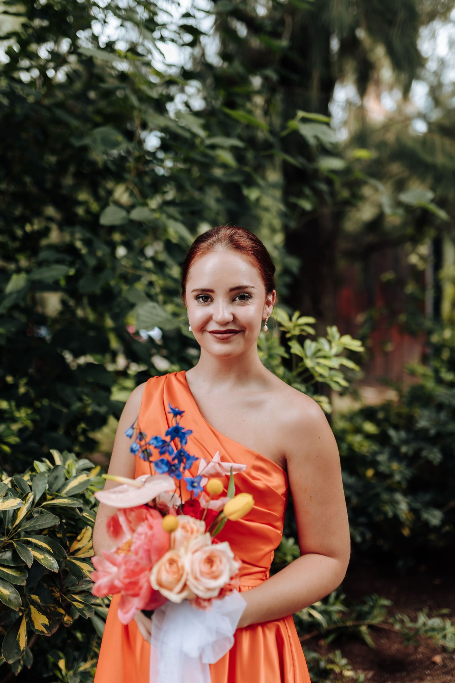 A bridesmaid in a bright coral one-shoulder dress holds a colorful bouquet against a lush garden backdrop.