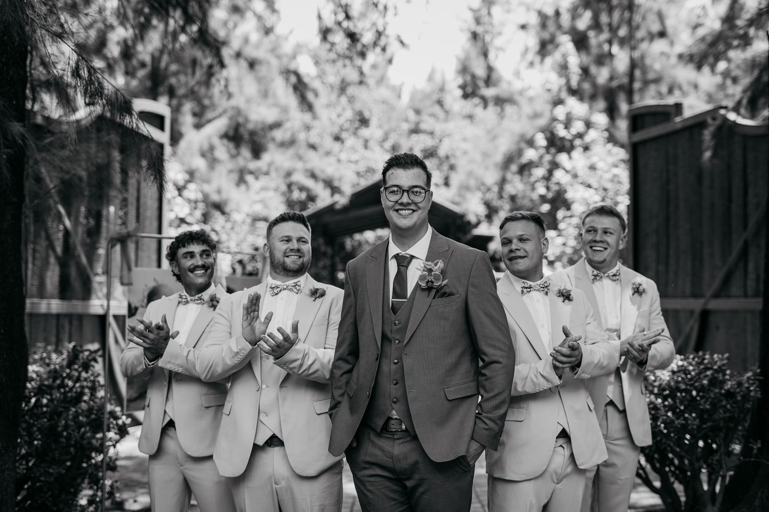 A joyful group of groomsmen in matching suits pose and laugh together during an outdoor wedding celebration.