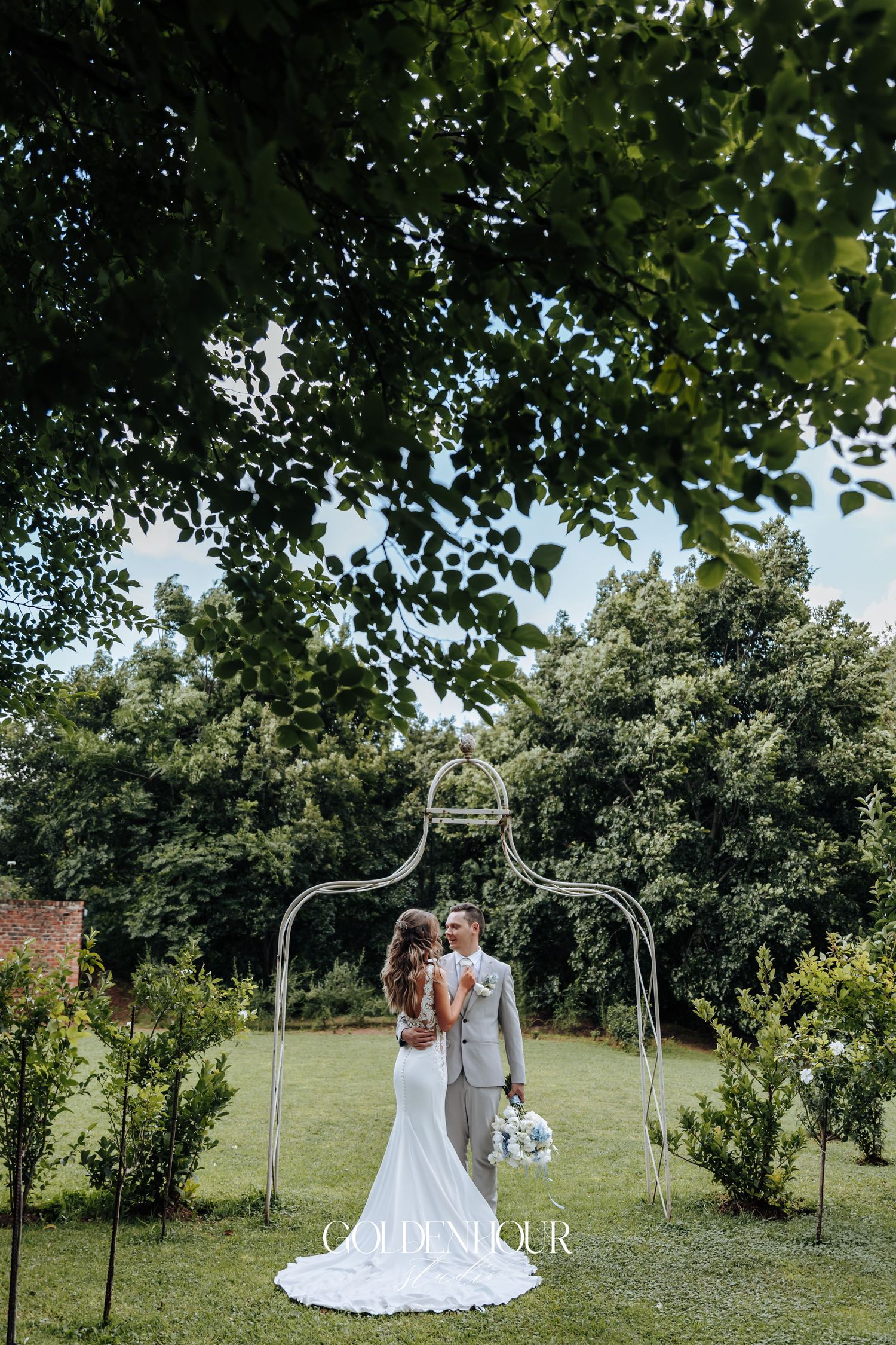 A wedding couple poses under a decorative metal arch in a lush garden setting.