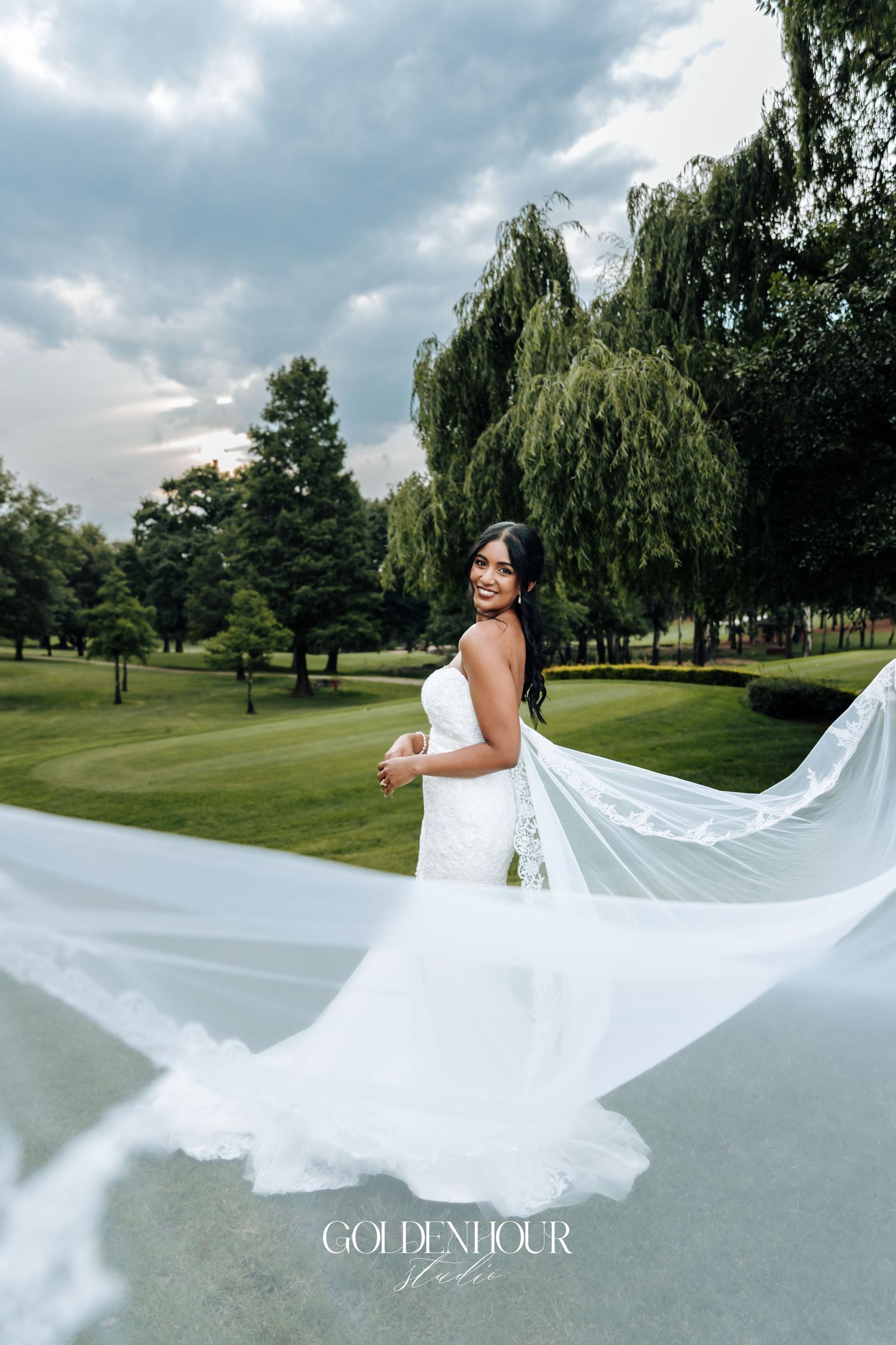 A bride in a flowing white dress stands on a golf course as her long veil billows dramatically in the wind.