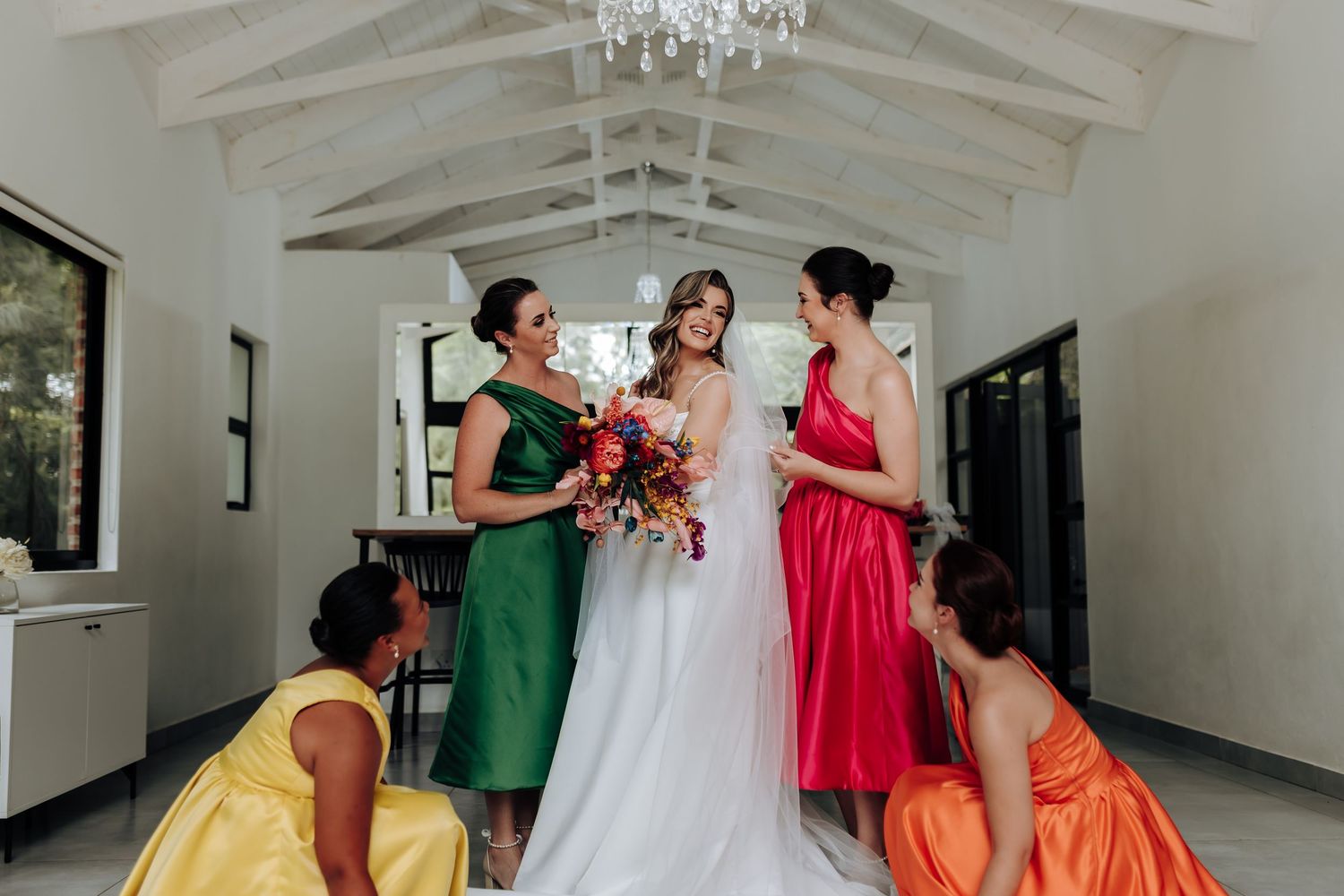 Wedding party in colorful dresses gathered around bride in white gown under vaulted ceiling.