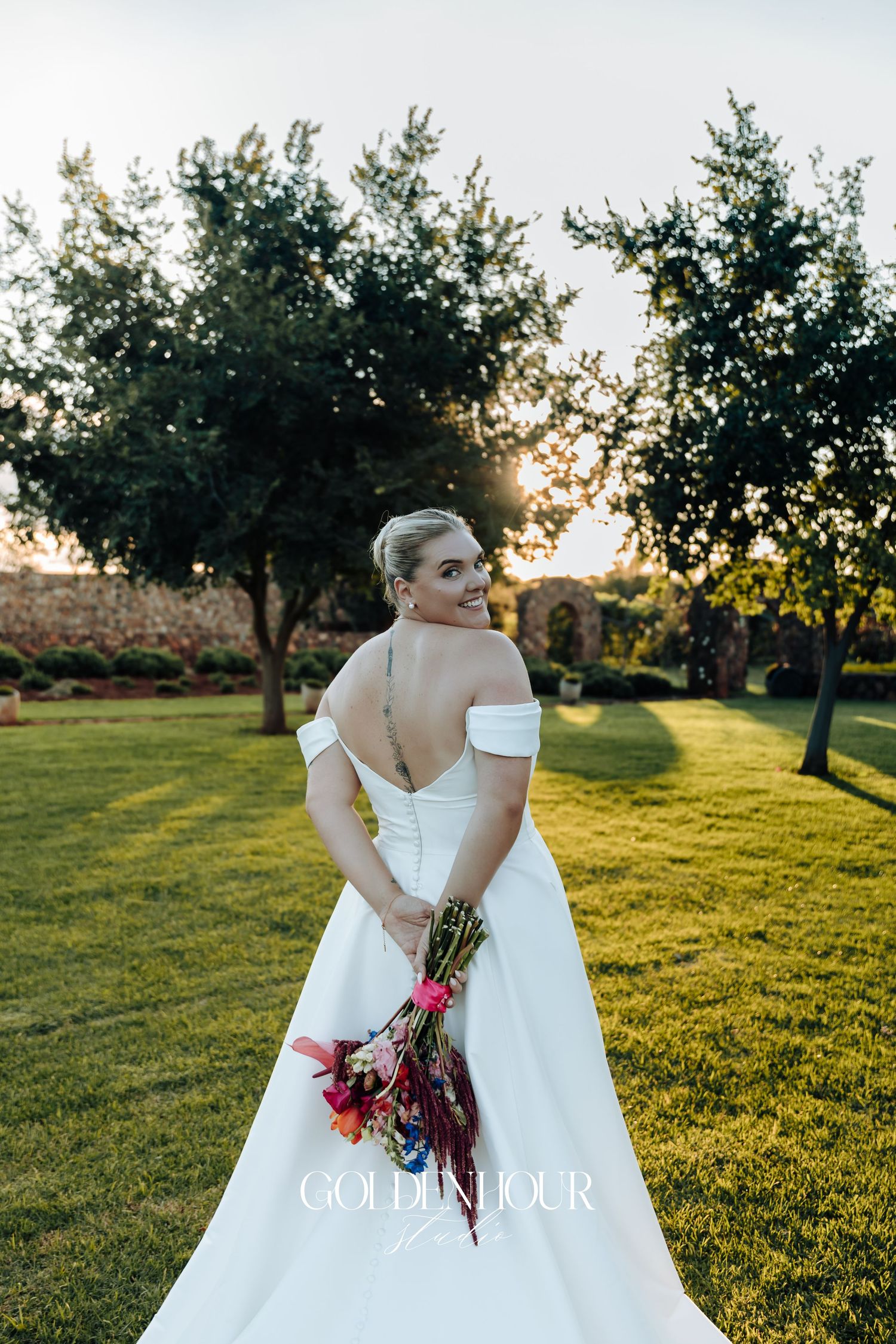A bride in a flowing white wedding dress holds a burgundy bouquet in a sunlit orchard during golden hour.