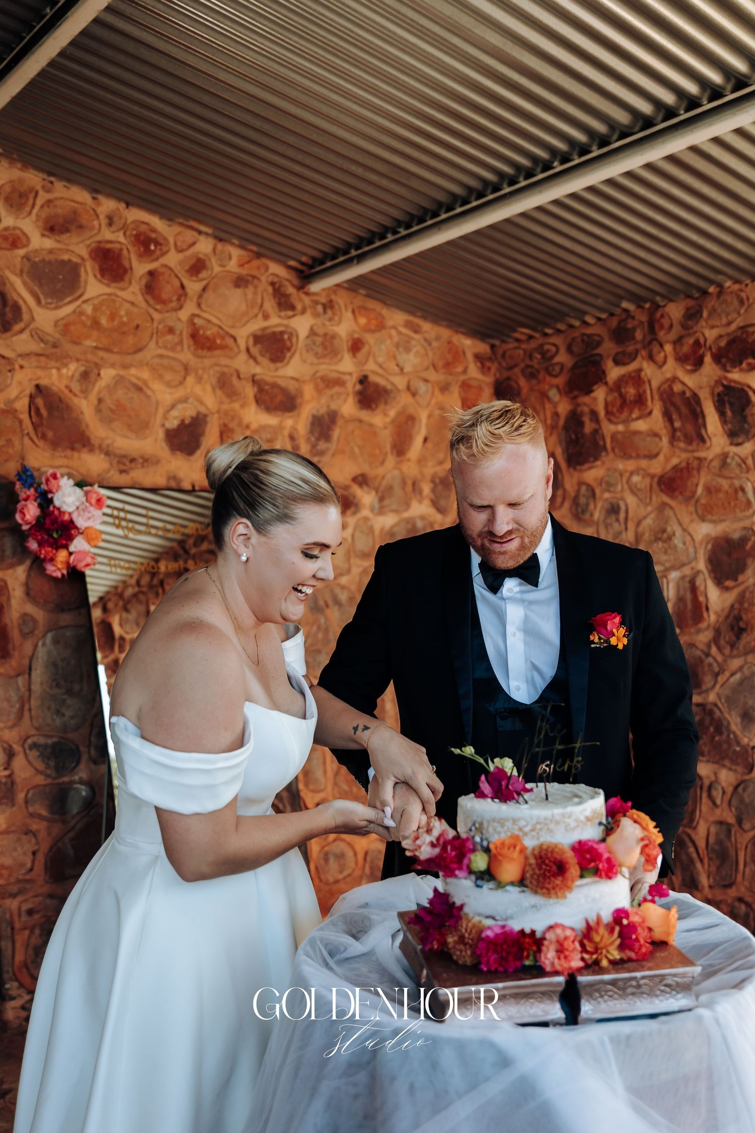 A couple shares a joyful moment cutting their wedding cake together at a rustic stone venue.