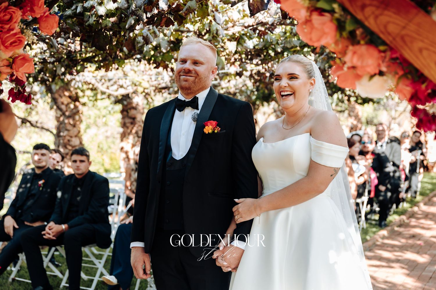 A joyful bride in a white off-shoulder gown walks down an outdoor aisle decorated with red flowers.