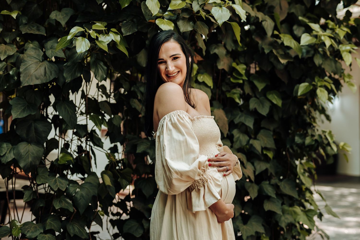 A woman in an elegant cream-colored dress poses in front of lush green ivy-covered walls.