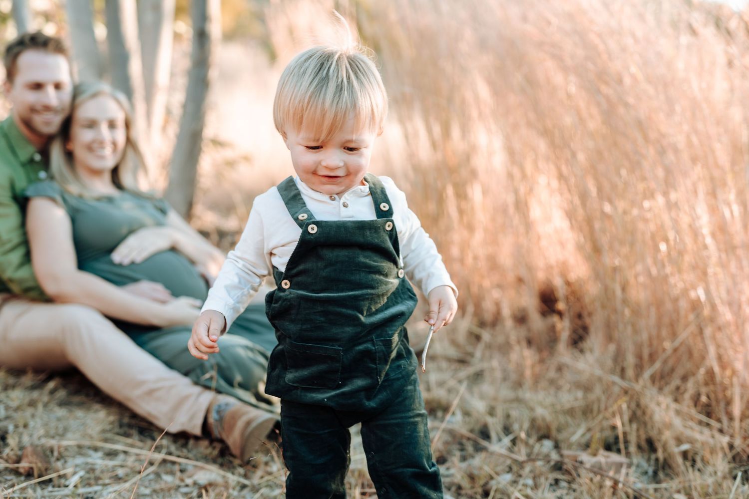 A toddler in green overalls stands in tall autumn grass while their parents sit in the background.