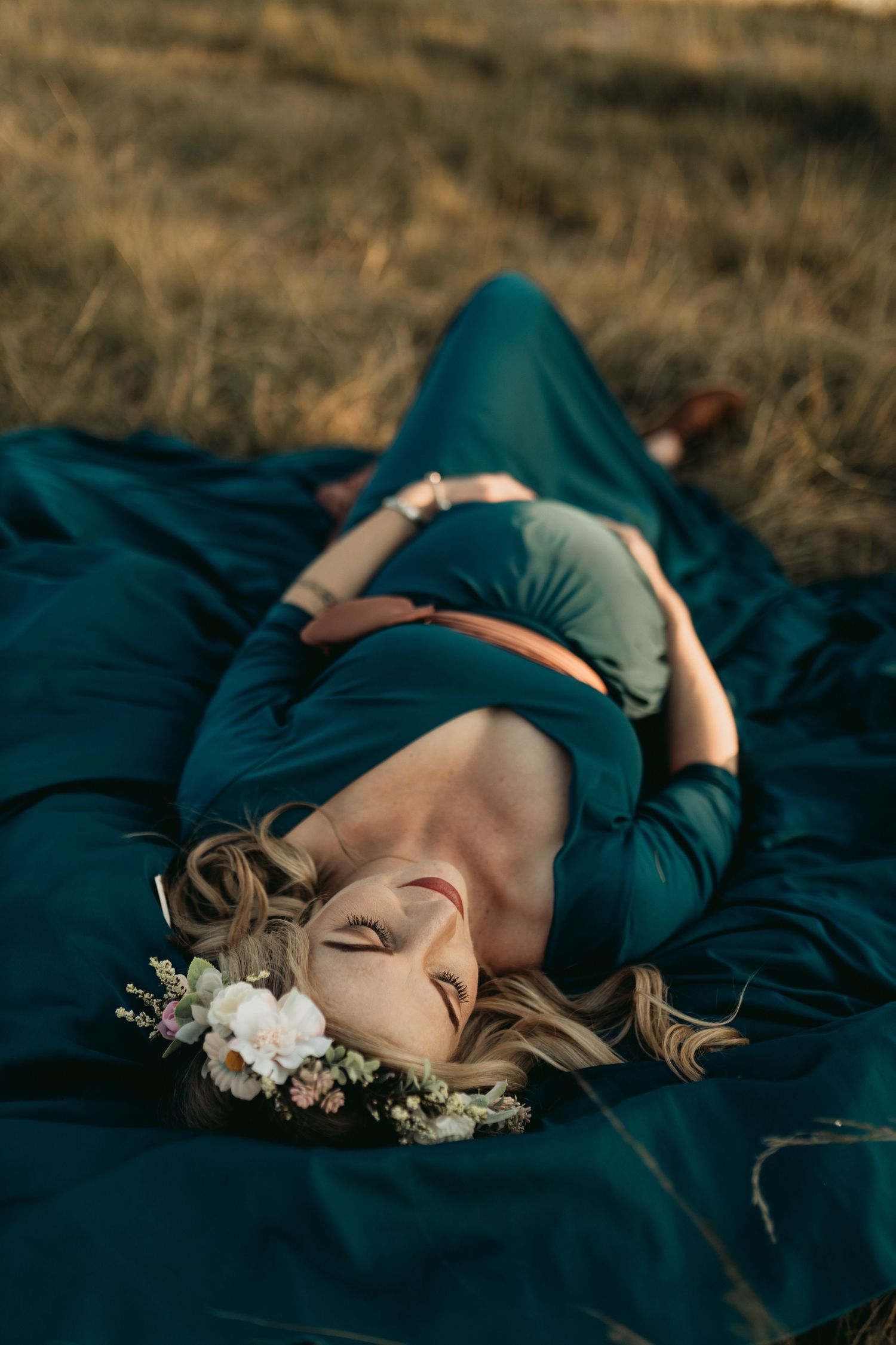 A woman in a teal dress reclines peacefully on a dark blanket with white flowers in her hair.