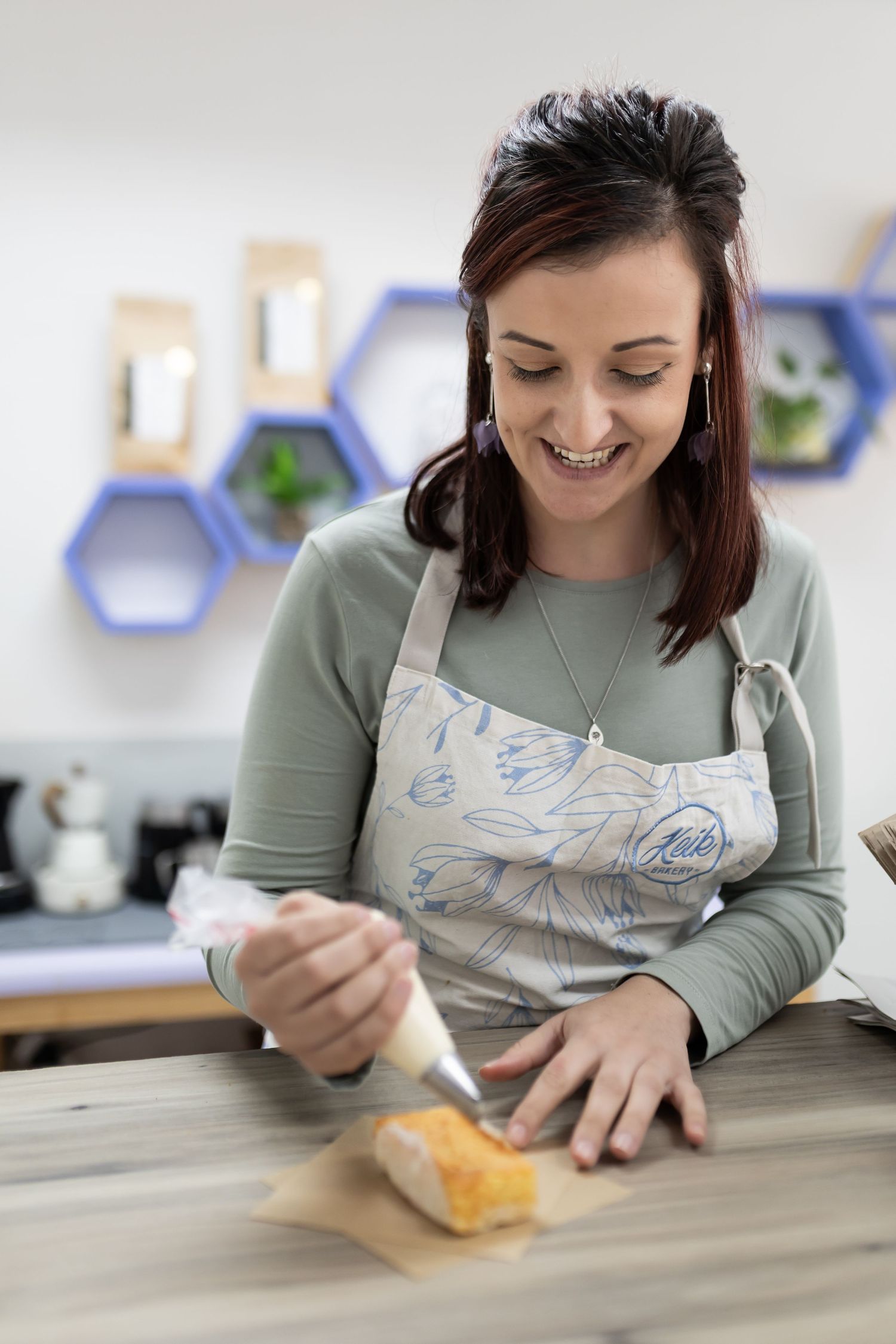 A person in a green shirt and apron writes on bubble mailer packaging at a desk with hexagonal wall shelves.
