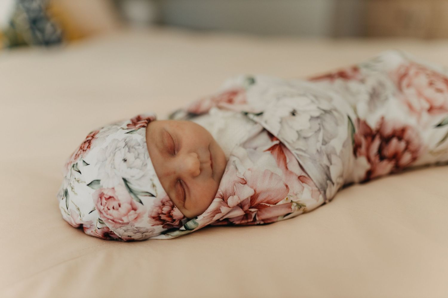 Newborn baby swaddled in floral print wrap sleeping peacefully on cream colored bedding.