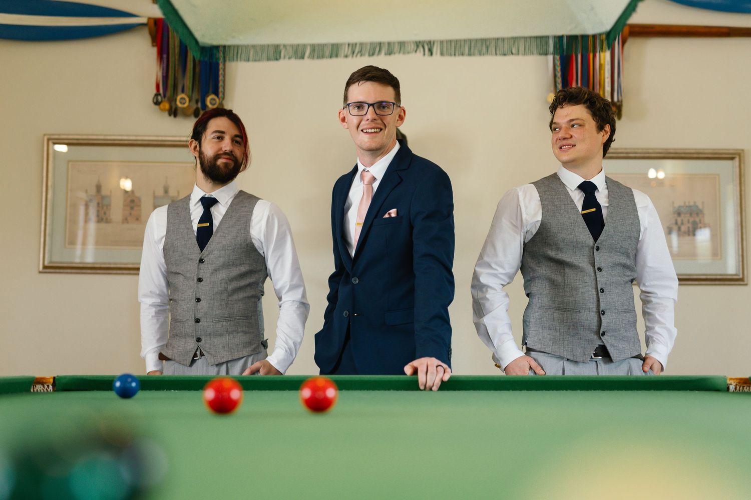 Groomsmen in formal suits gather around a green pool table during a wedding celebration.