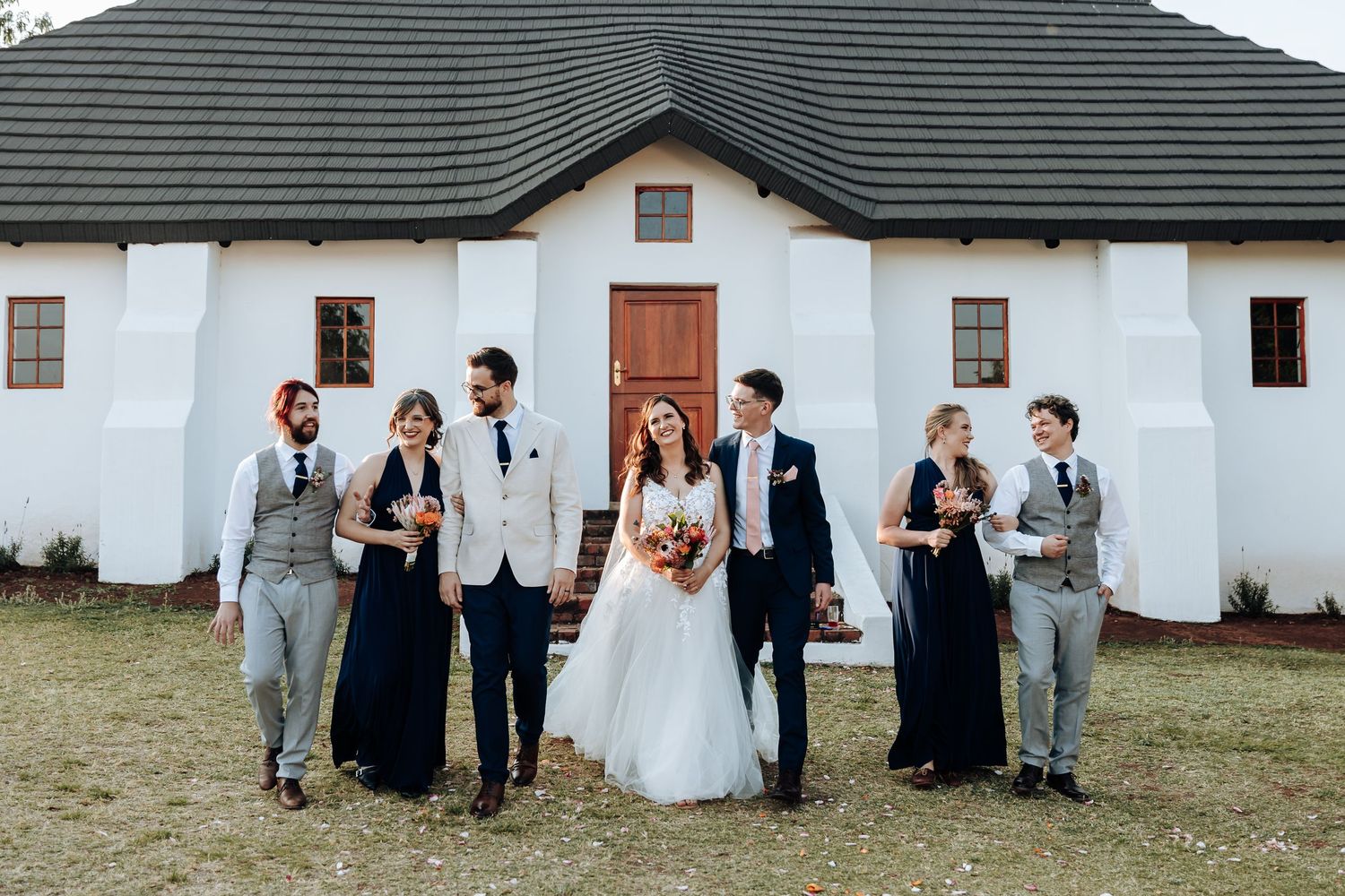 Wedding party members pose against a rustic white wall and green tiled roof building in a candid outdoor setting.