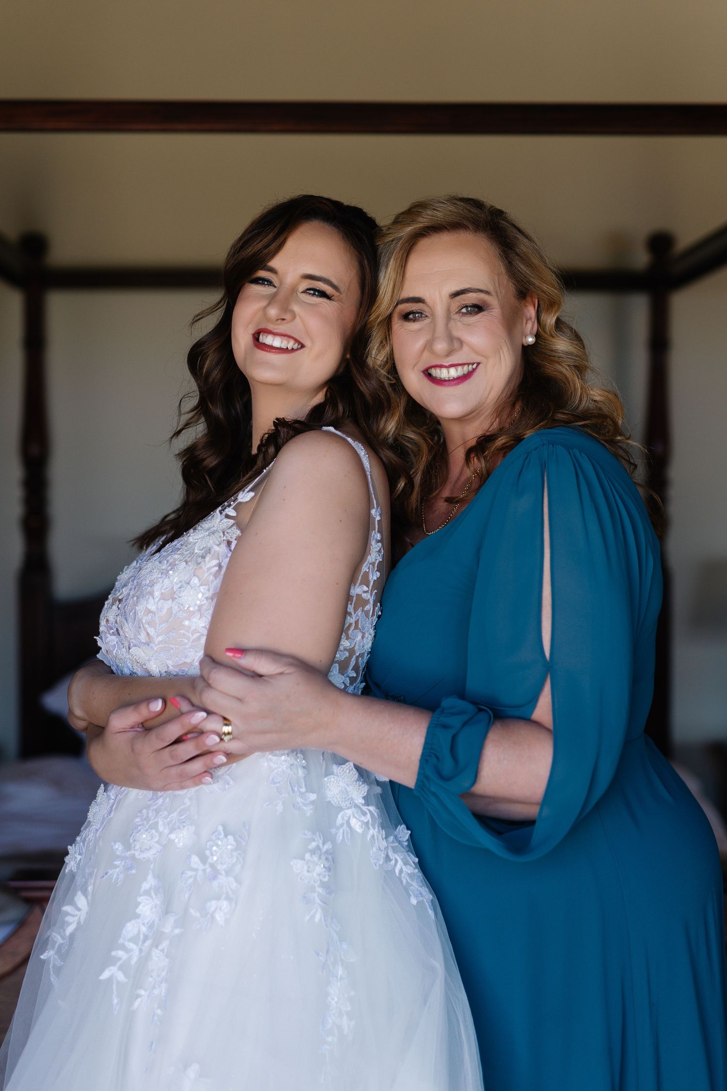 Two women embrace and smile warmly, one wearing a white lace wedding dress and the other in a blue dress.