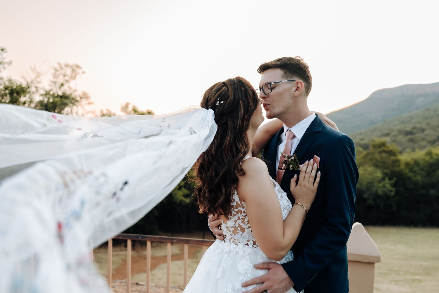 A romantic wedding couple embraces outdoors with a flowing veil catching the breeze against a mountain backdrop at sunset.