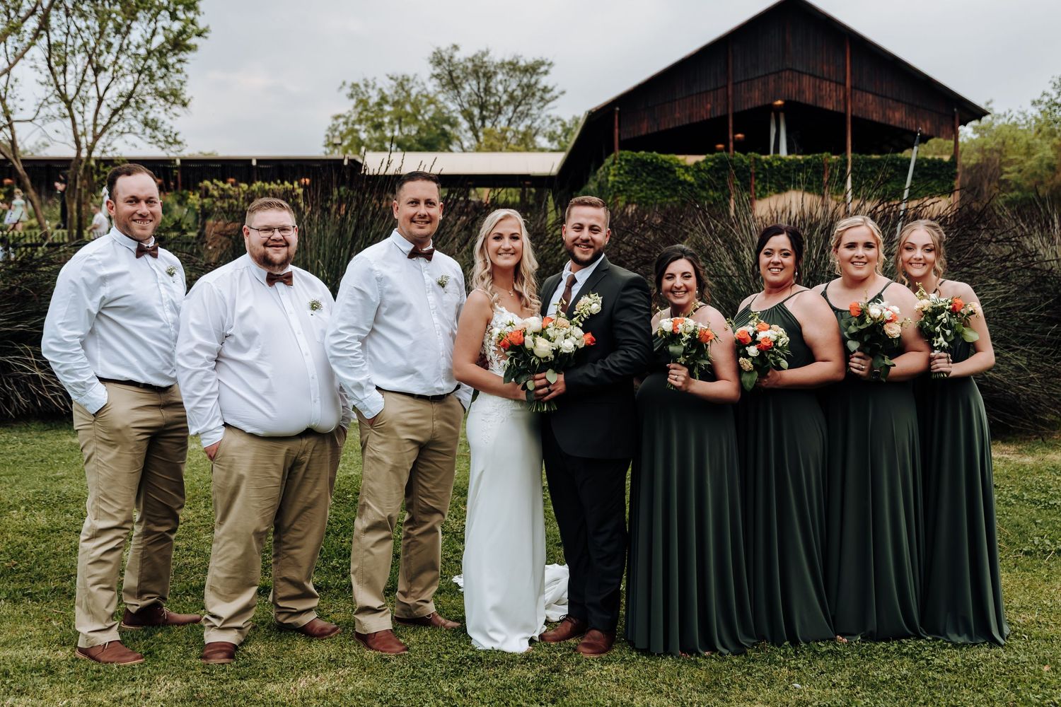 Wedding party poses outdoors with groomsmen in khakis and bridesmaids in dark green dresses by rustic barn.