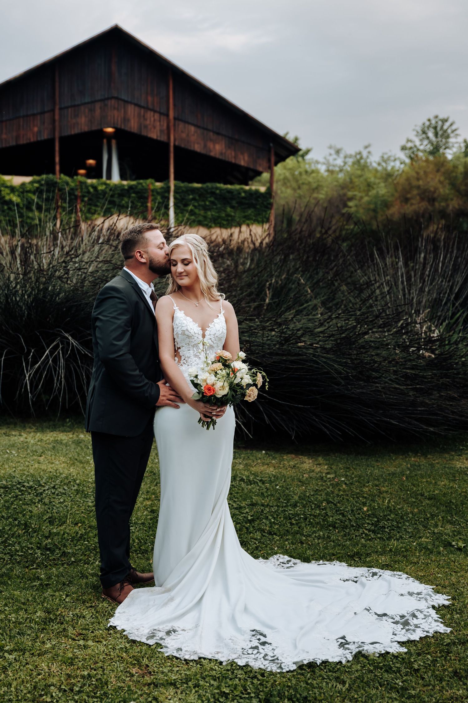 A romantic couple in wedding attire embraces on a green lawn with a rustic barn in the background.