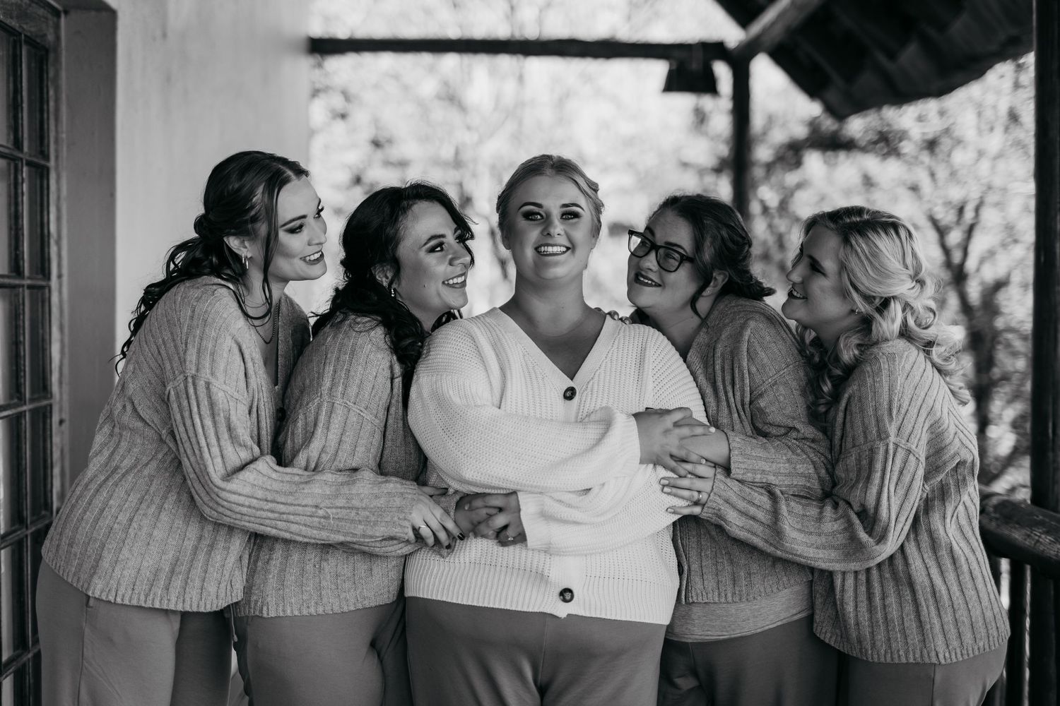 Bride surrounded by bridesmaids in matching sweaters sharing a joyful group hug in black and white photo.