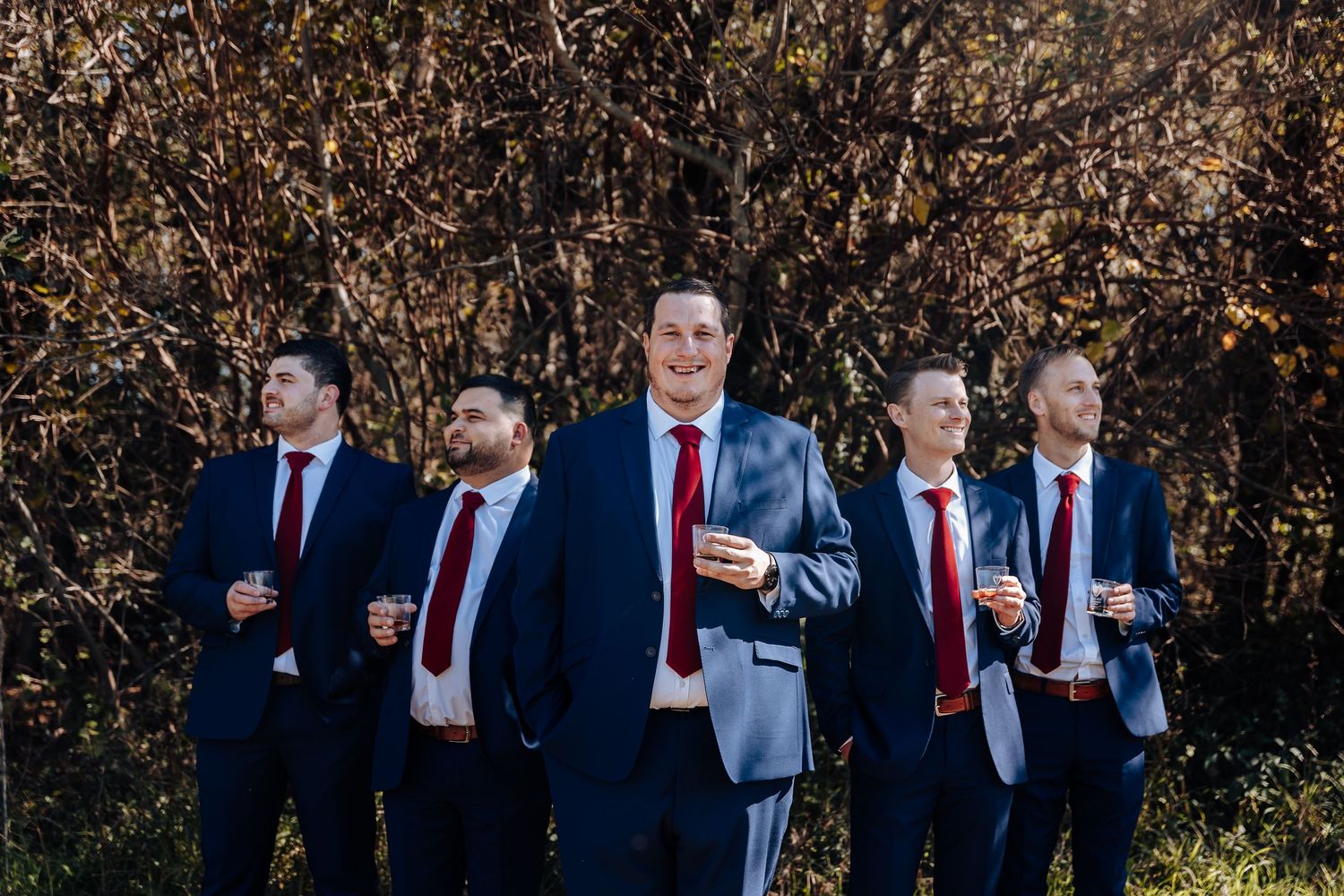 Five groomsmen in navy suits and red ties pose outdoors among autumn foliage for a wedding party photo.