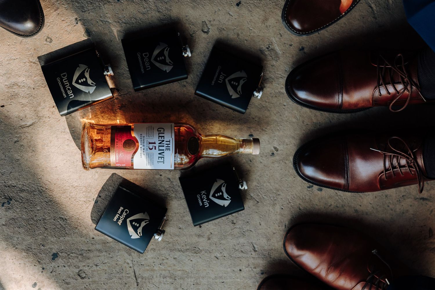 Overhead view of whiskey bottle, black flask gift sets, and brown leather dress shoes on concrete floor.