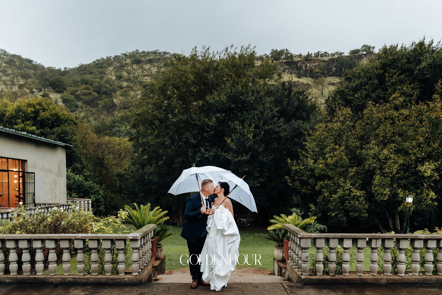 Romantic couple kissing under a white umbrella in lush garden with mountain backdrop.
