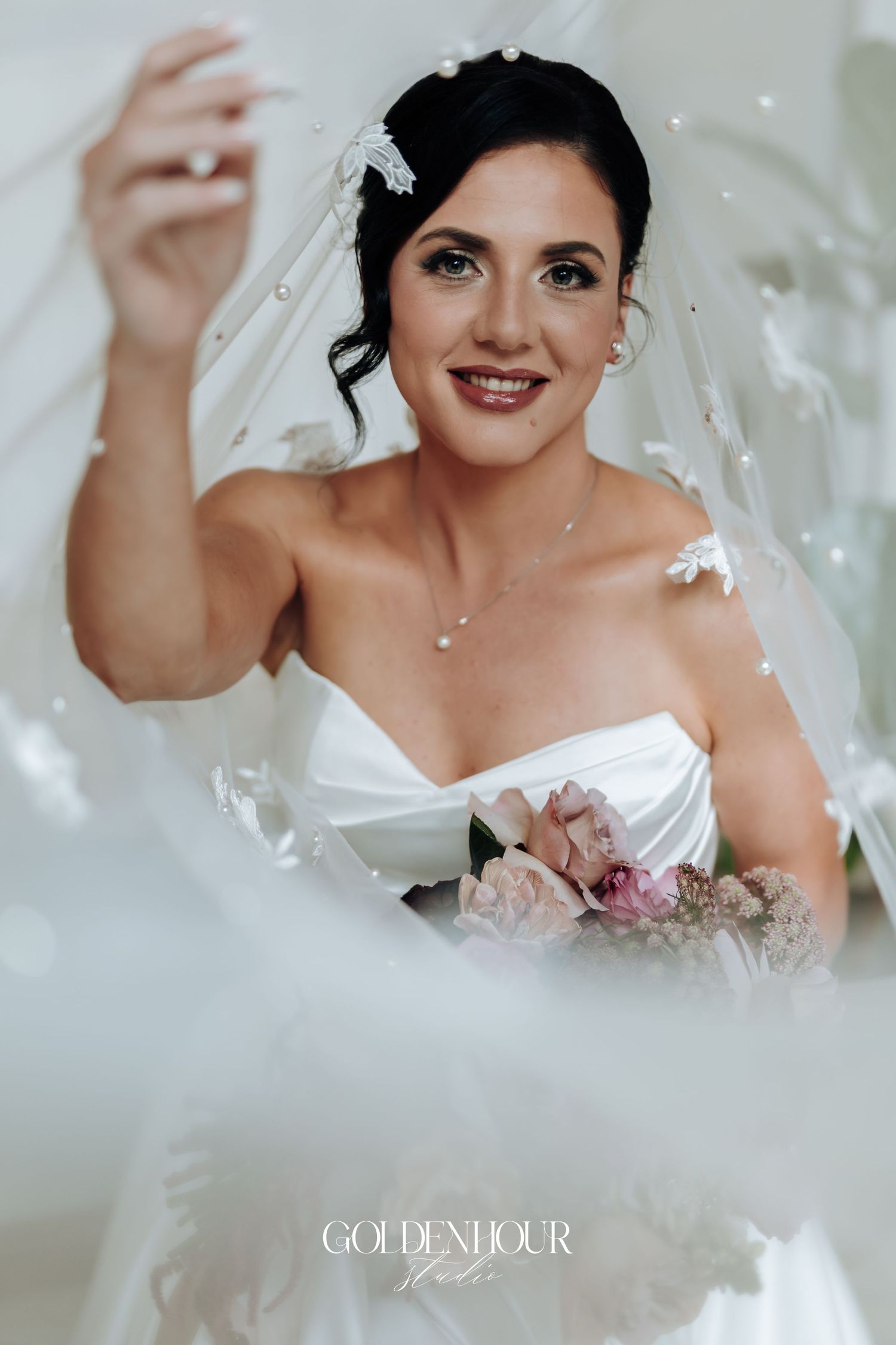 Beautiful bride in strapless white gown holding pink bouquet beneath pearl-adorned wedding veil.
