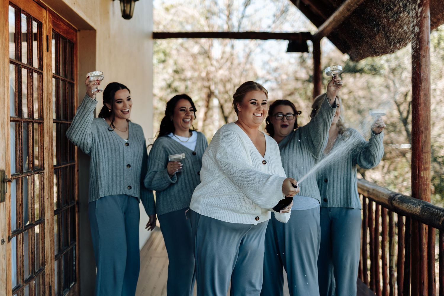 Bridesmaids in blue pajamas celebrate with champagne spray on a wooden porch during a fun bridal party morning.