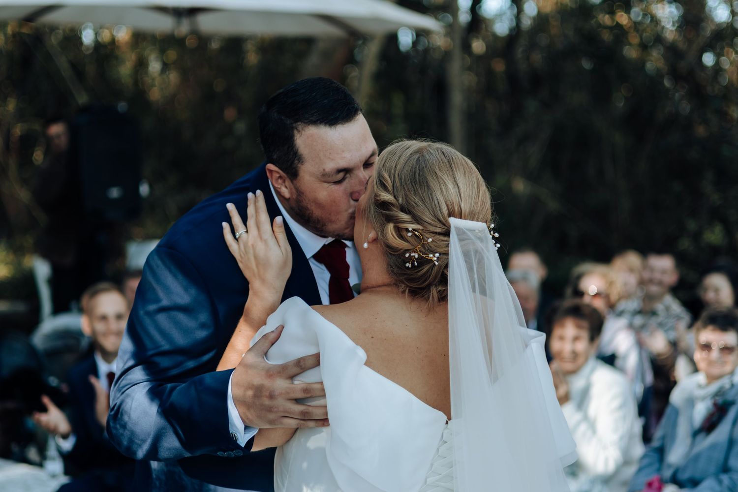 A bride and groom share their first kiss at an outdoor wedding ceremony surrounded by applauding guests.