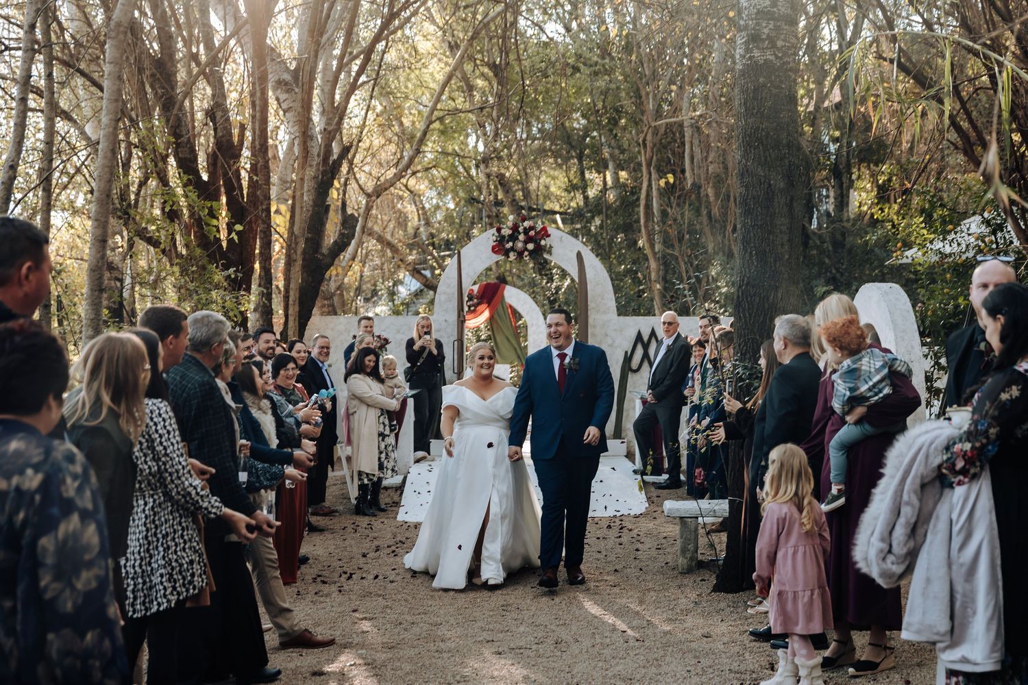 Bride and groom walk down outdoor aisle surrounded by guests and trees at a romantic woodland wedding ceremony.
