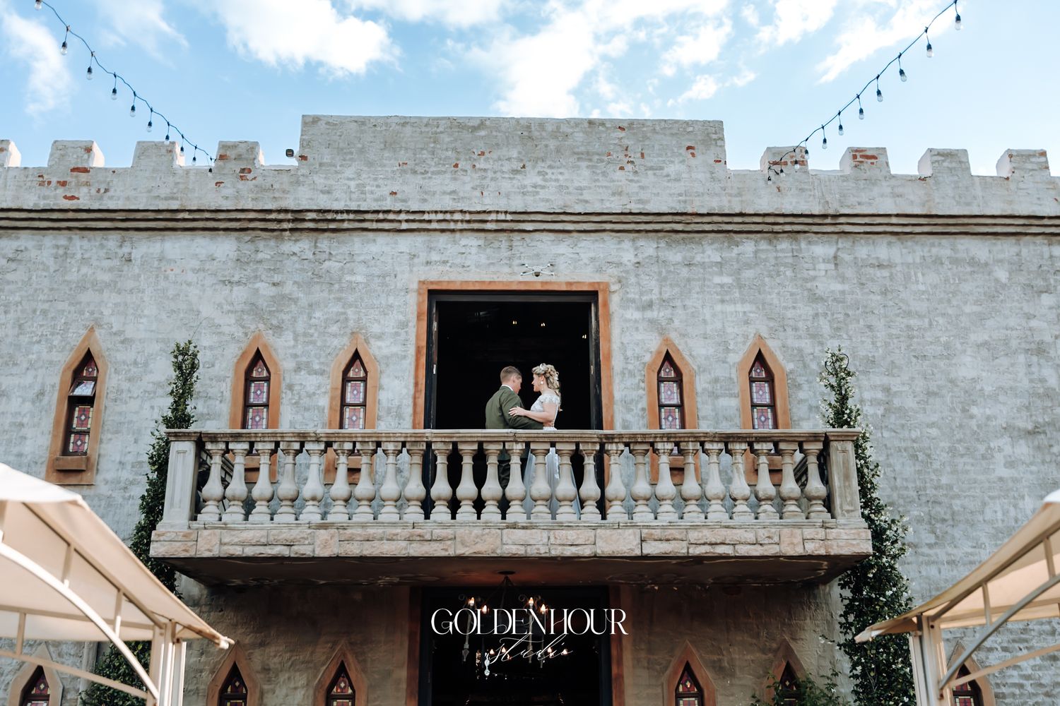 Bride and groom embracing on castle balcony with string lights and gothic arched windows below.