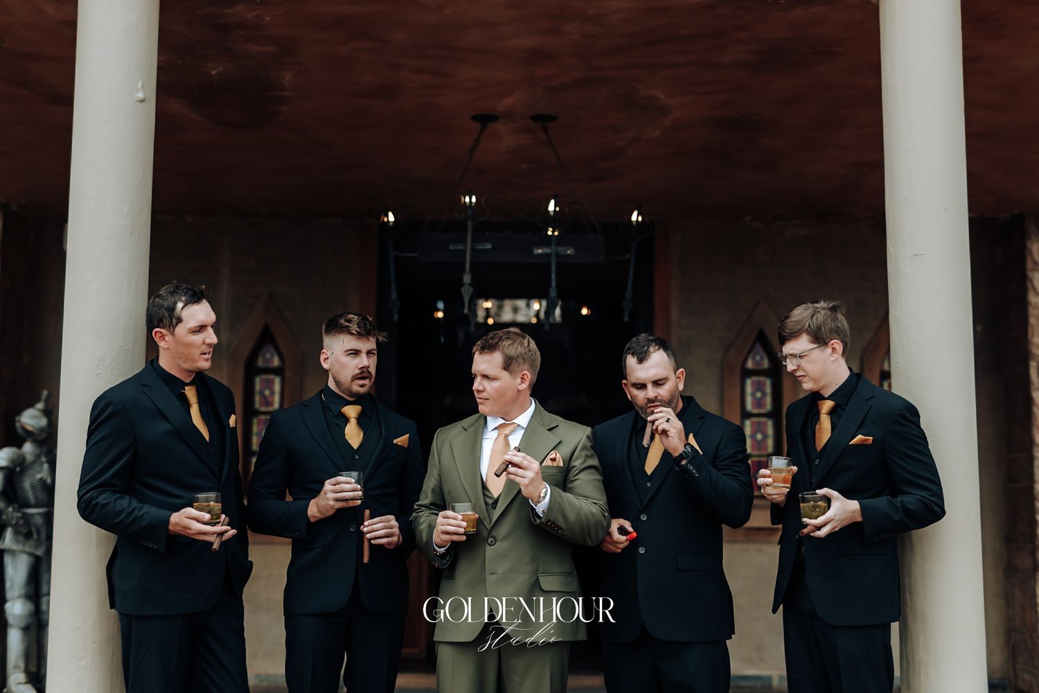 Groom in tan suit stands with four groomsmen in navy suits and gold ties, holding drinks at an elegant wedding venue.