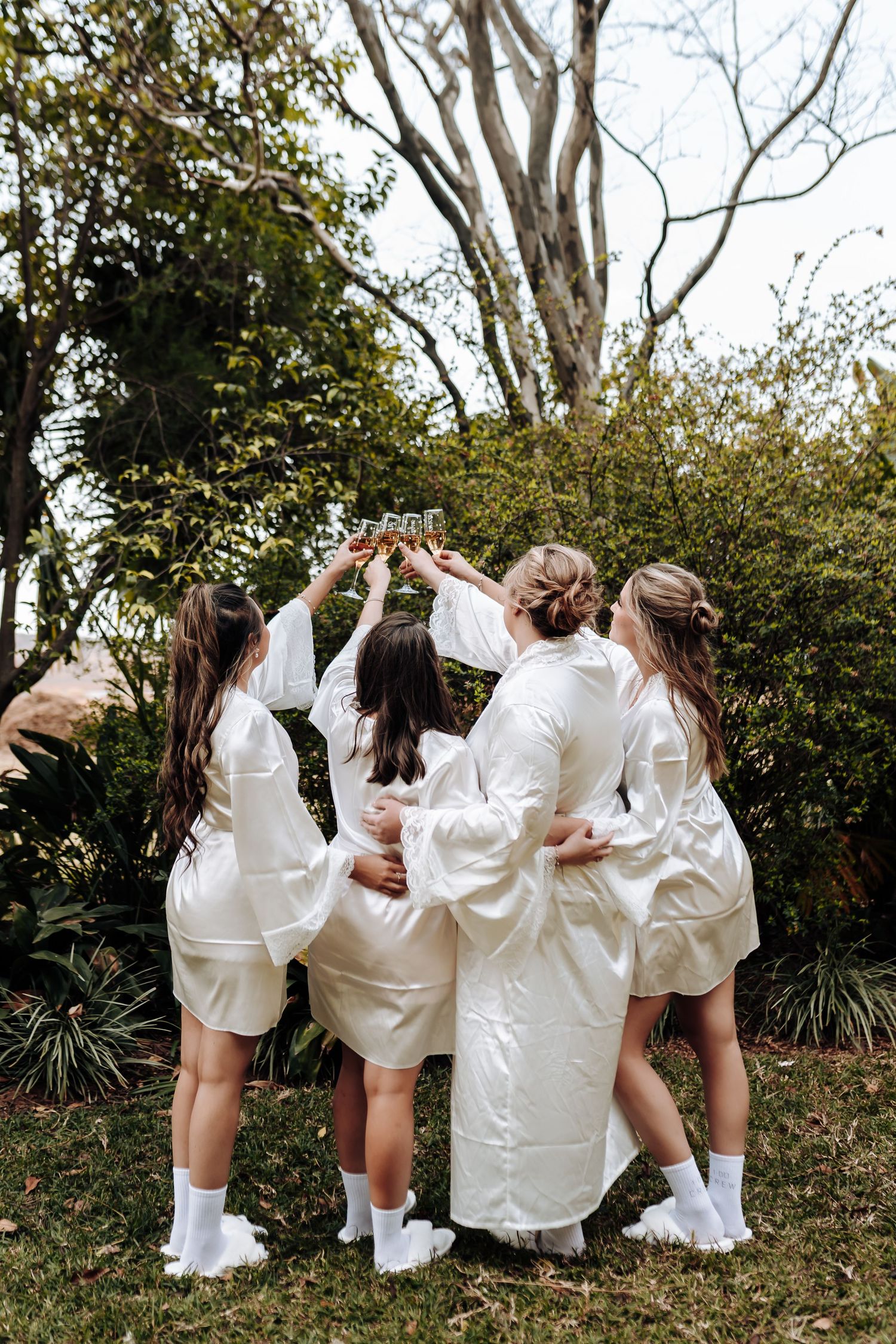 A group in matching white satin robes and sneakers huddles together outdoors while raising their hands in celebration.