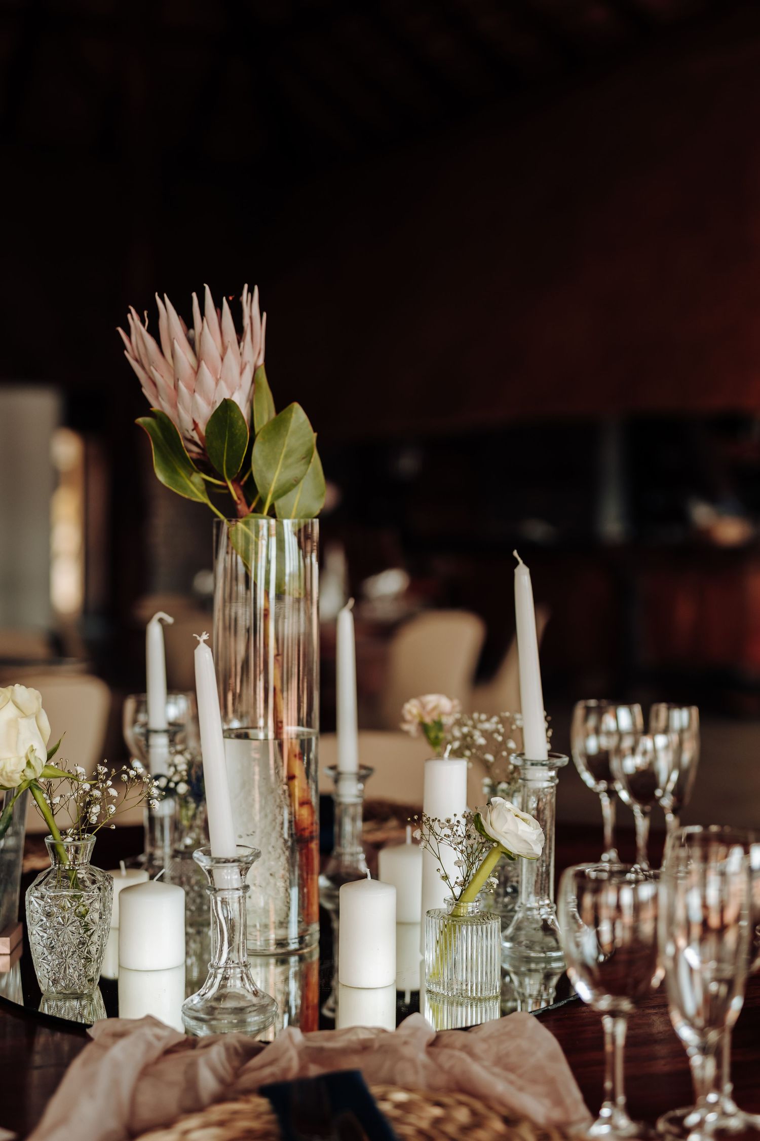 Elegant wedding table centerpiece with pink protea flower, white candles and crystal glassware on dark background.