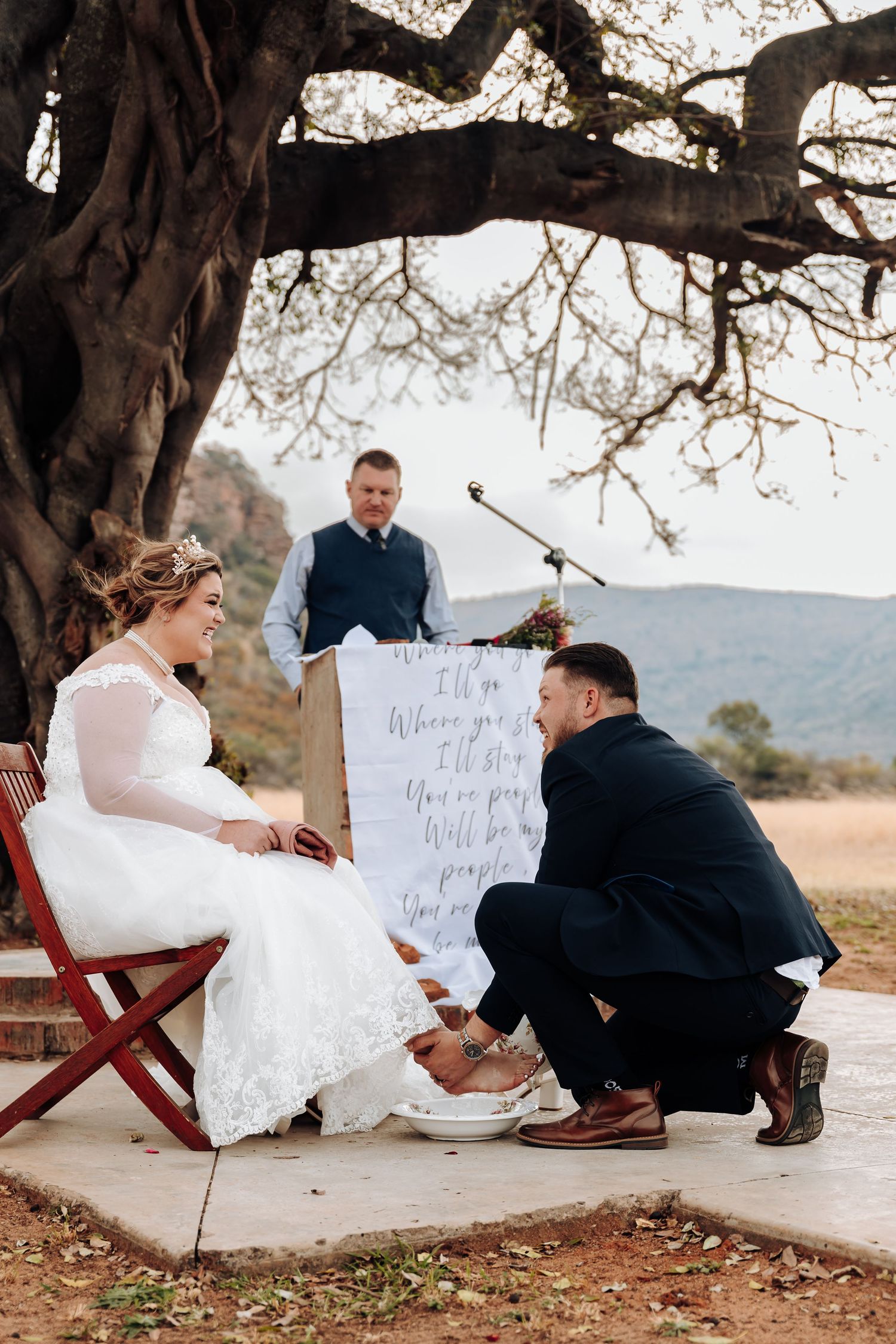 A wedding ceremony outdoors under a large tree with a couple in formal attire on a concrete platform.