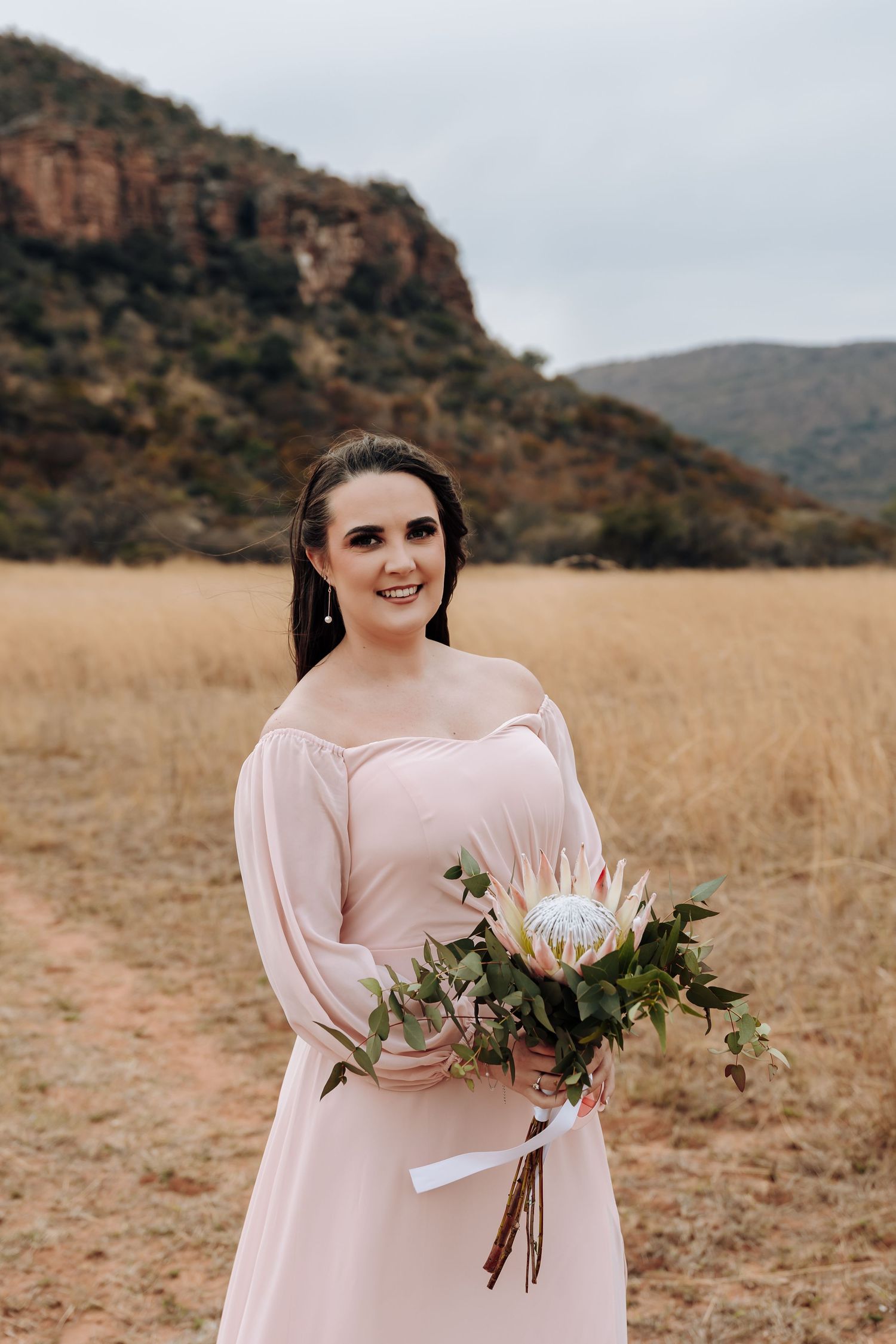 Figure in a blush pink dress holds a protea bouquet against a backdrop of mountains and dry grassland.