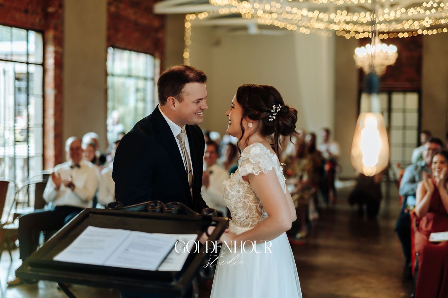 Bride and groom smiling at each other during wedding ceremony with guests and chandelier in background.