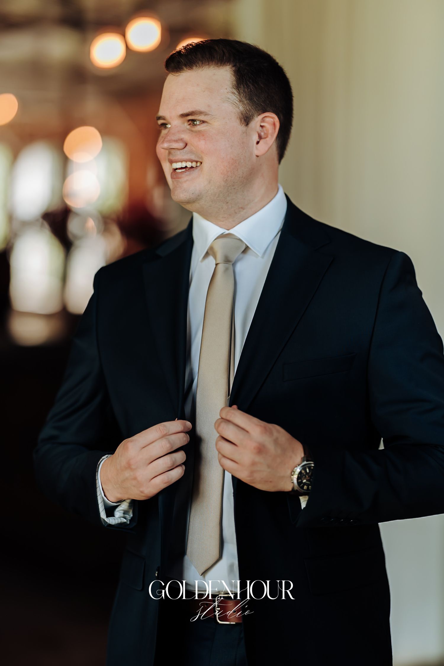 Smiling groom in dark navy suit adjusting his beige tie before the wedding ceremony.