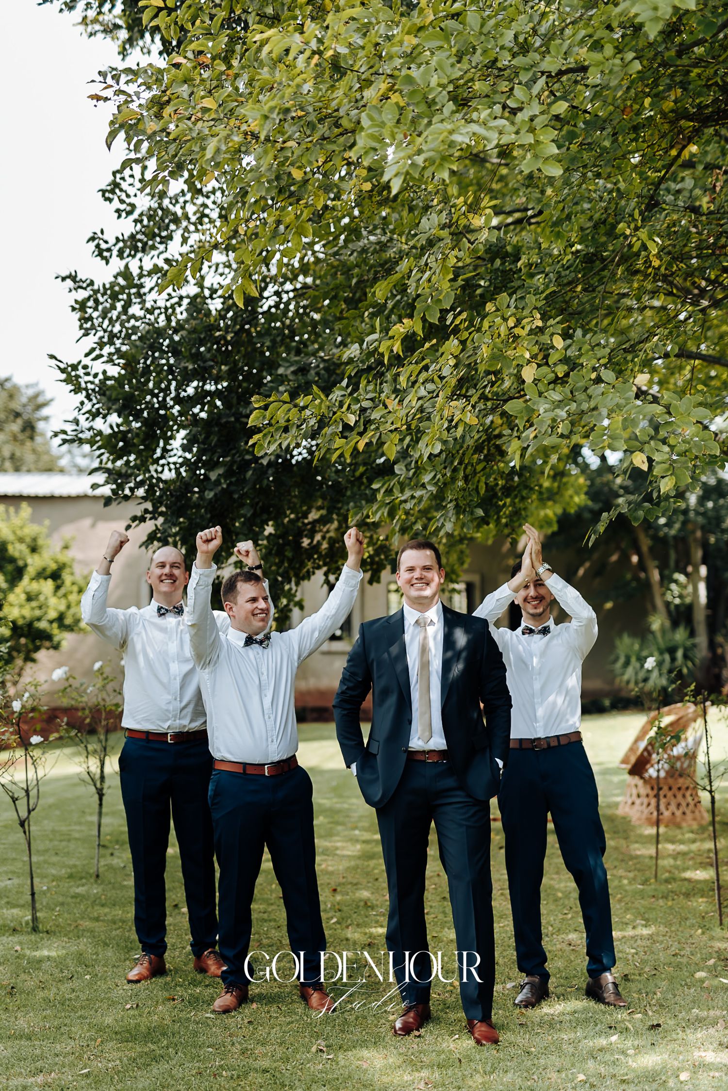 Groom and groomsmen celebrating outdoors in navy suits and white shirts under a green tree.